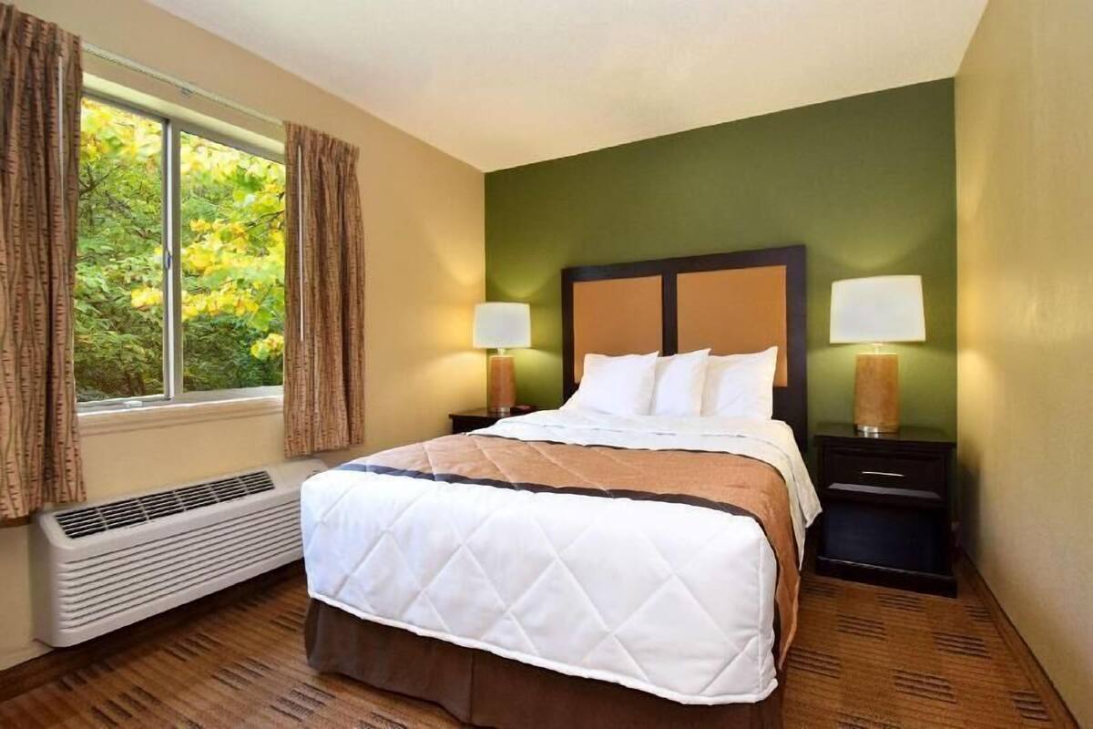 A hotel room with a bed, two bedside tables with lamps, a window with curtains, and a wall heater.