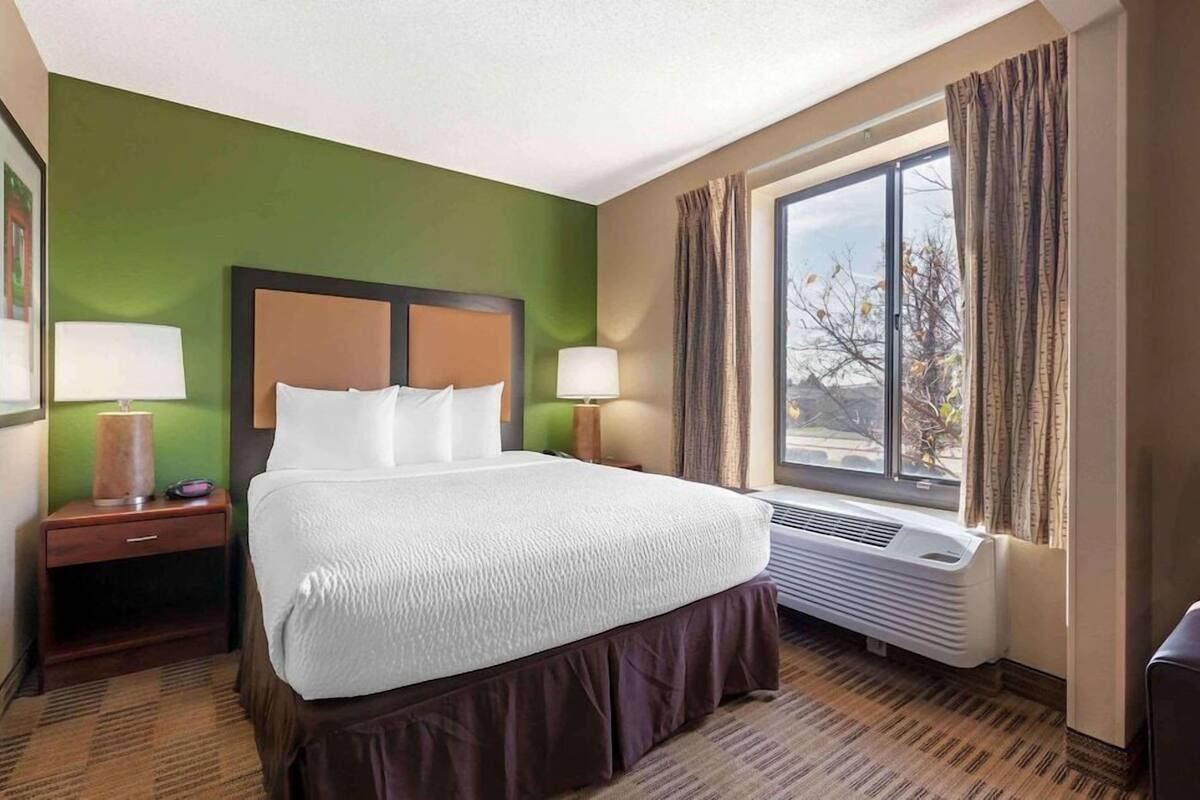 Una camera d'hotel con un letto grande, due comodini con lampade, una finestra con tende e un condizionatore d'aria a parete.