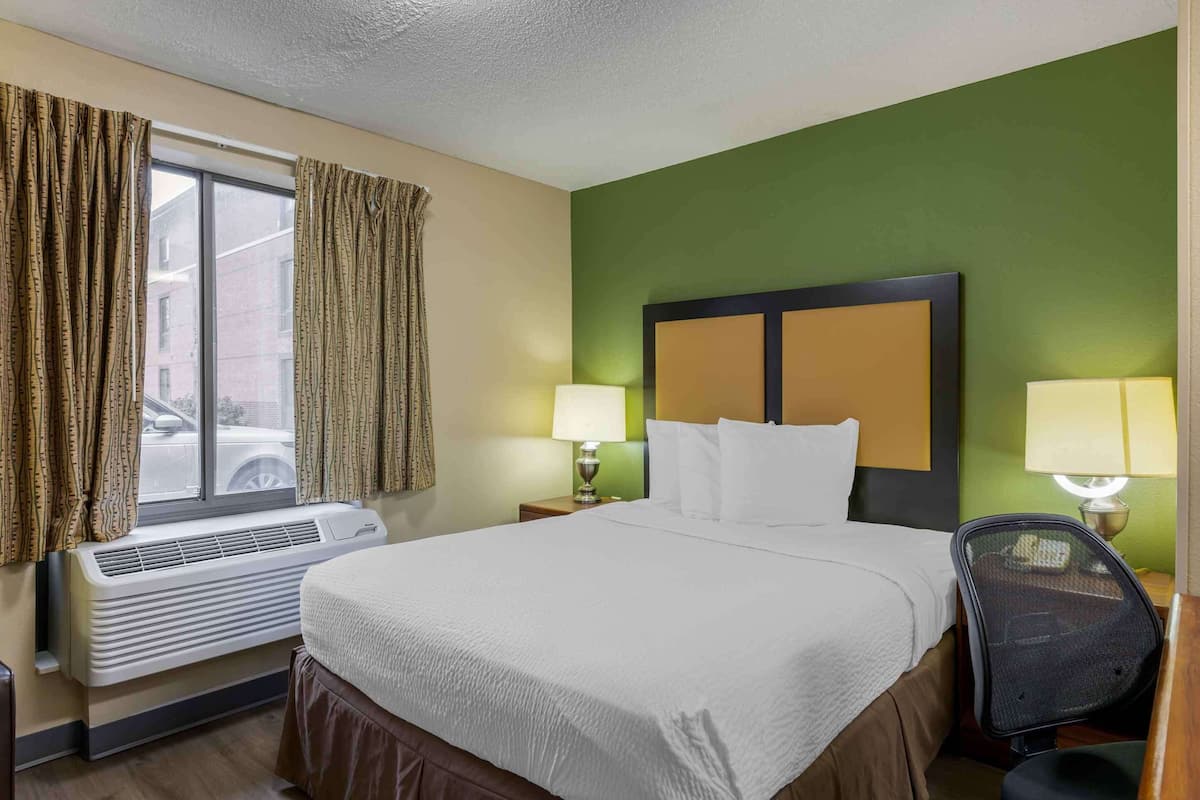 A hotel room with a bed, two bedside lamps, a window with curtains, an air conditioner, and a chair.