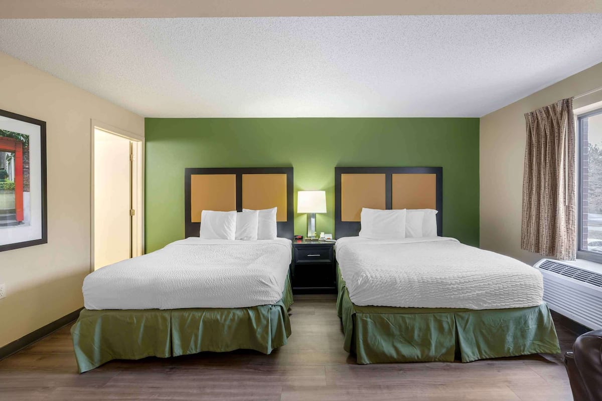 A hotel room with two beds, a nightstand with a lamp, a window with curtains, and a framed picture on the wall.