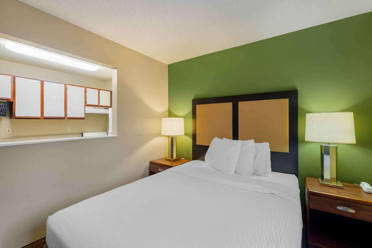 A hotel room with a bed, two bedside tables with lamps, a nightstand, a mirror, and a window with blinds.
