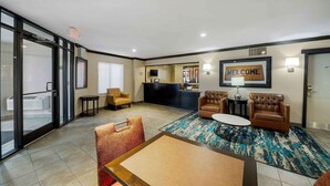 A modern hotel lobby with a reception desk, a 'WELCOME' sign, leather sofas, a coffee table, and a glass door leading to a balcony. - Extended Stay America Suites Oklahoma City Northwest (Oklahoma City)
