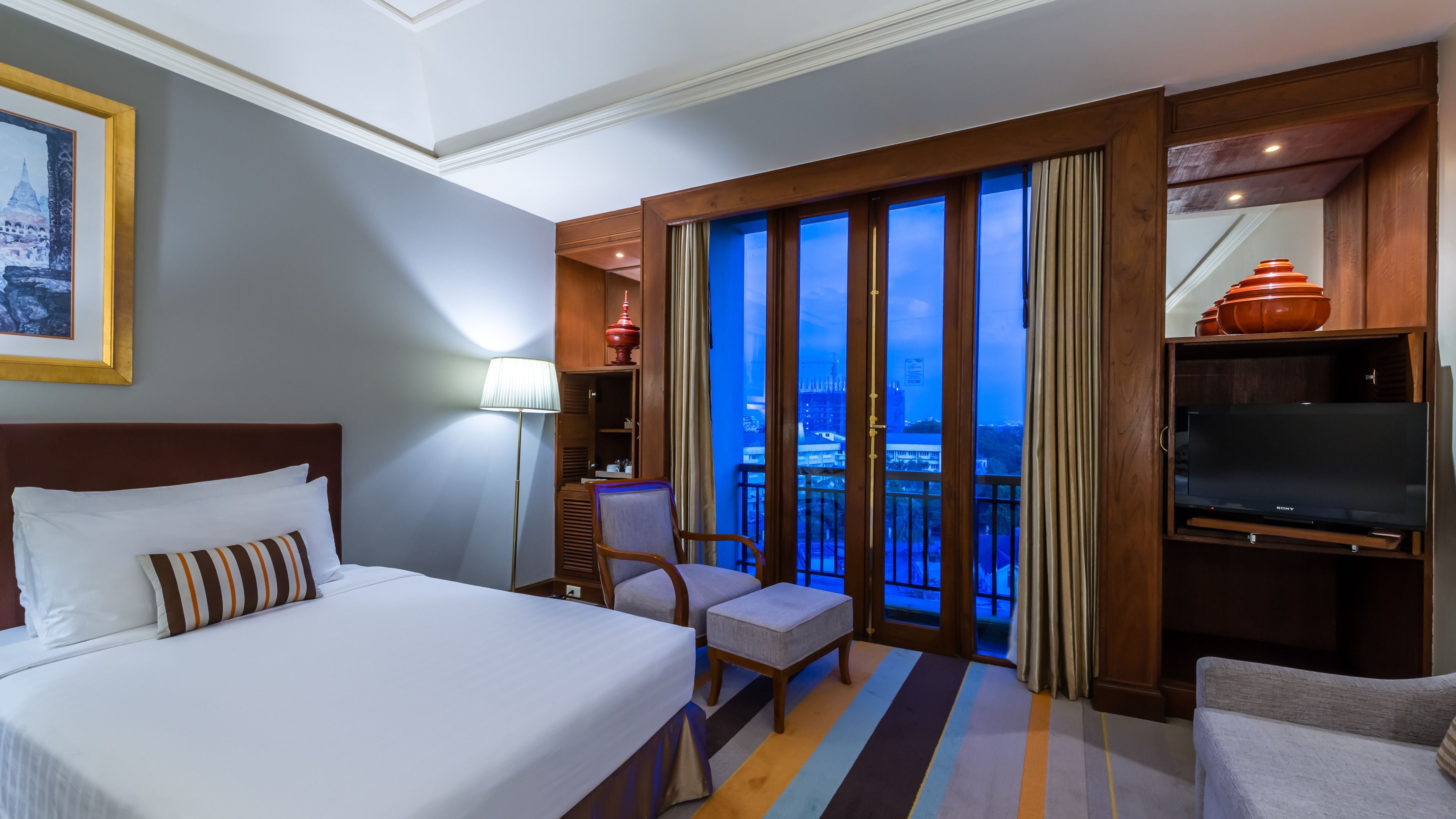 Habitación de hotel con una cama grande, una silla, un televisor y vistas a la ciudad desde un balcón.
