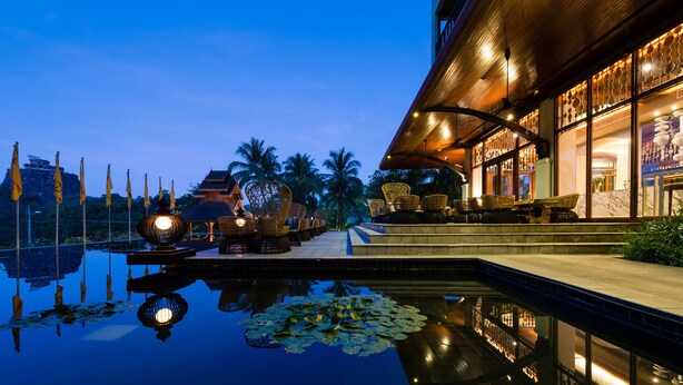 A serene evening pool with lotus leaves, a traditional building with a wooden facade, and a clear view of palm trees in the background. - Chatrium Hotel Royal Lake Yangon (Yangon)
