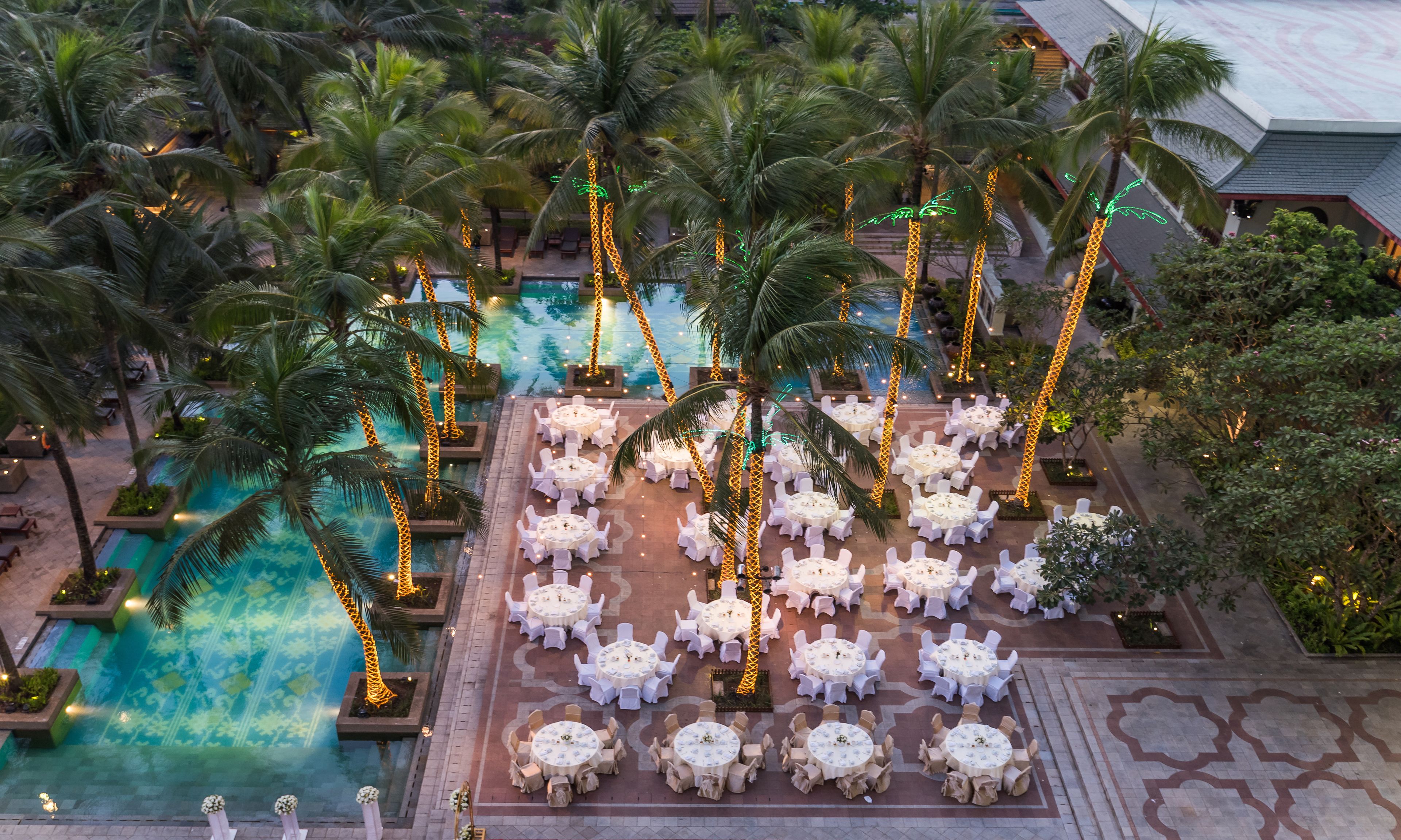An aerial view of a resort with a pool surrounded by palm trees and outdoor seating areas.