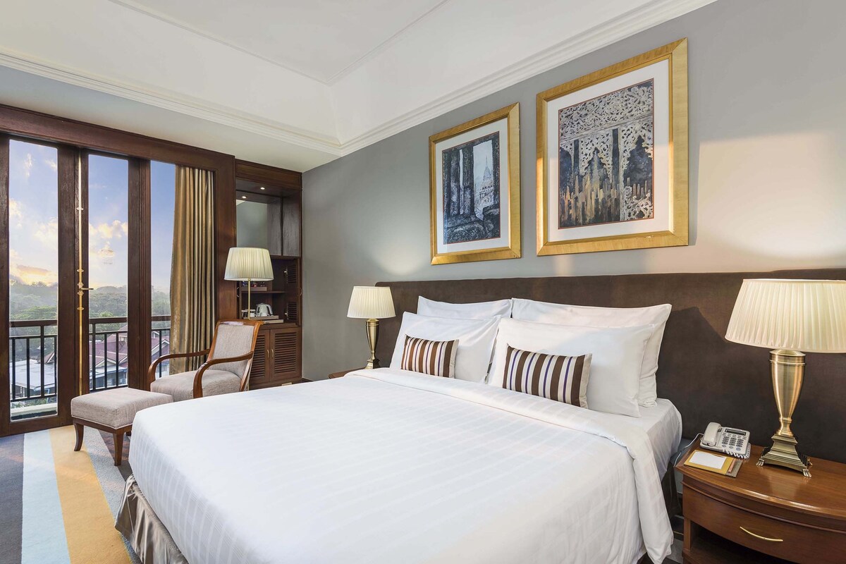 A hotel room with a large bed, two bedside lamps, a nightstand with a phone, a chair, a desk with a lamp, and two framed pictures on the wall.