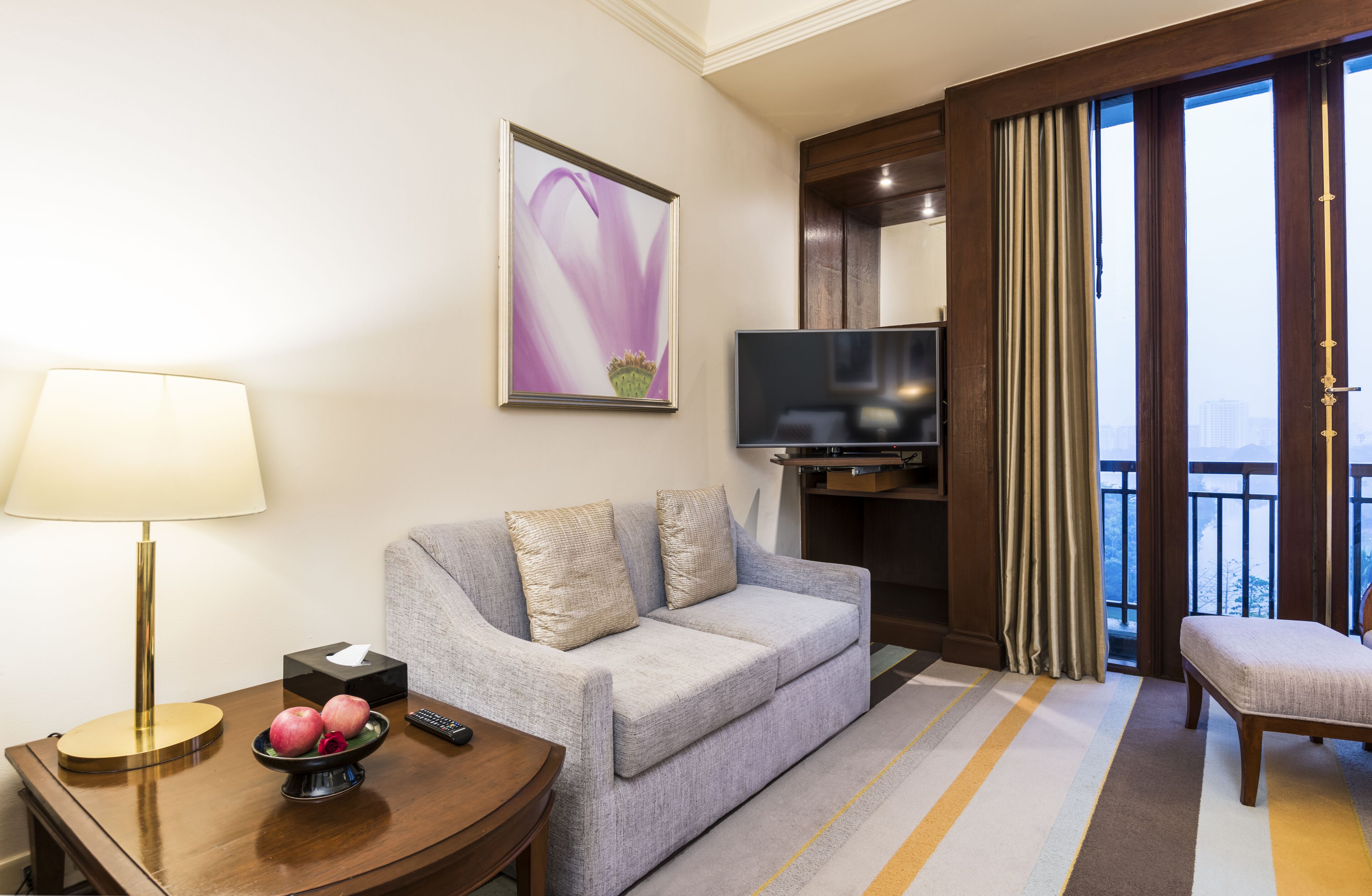 A hotel room with a grey sofa, a TV, a bedside table with a lamp, a bowl of fruit, and a window with curtains.