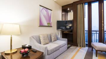 Habitación de hotel con un sofá gris, un televisor, una mesita de noche con lámpara, un cuenco de fruta y una ventana con cortinas.