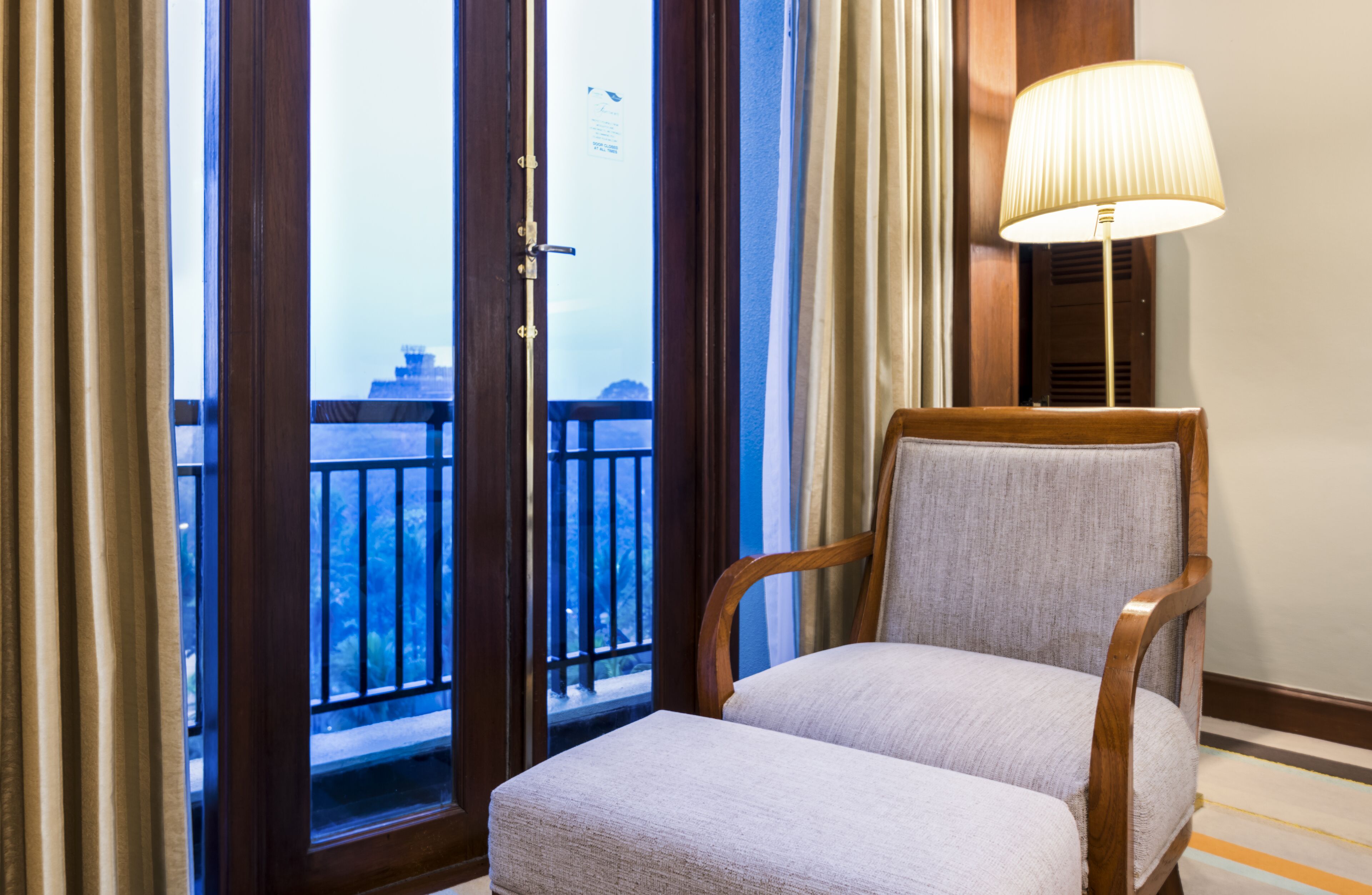 A room with a wooden chair, a lamp, and a view of a balcony with a railing.