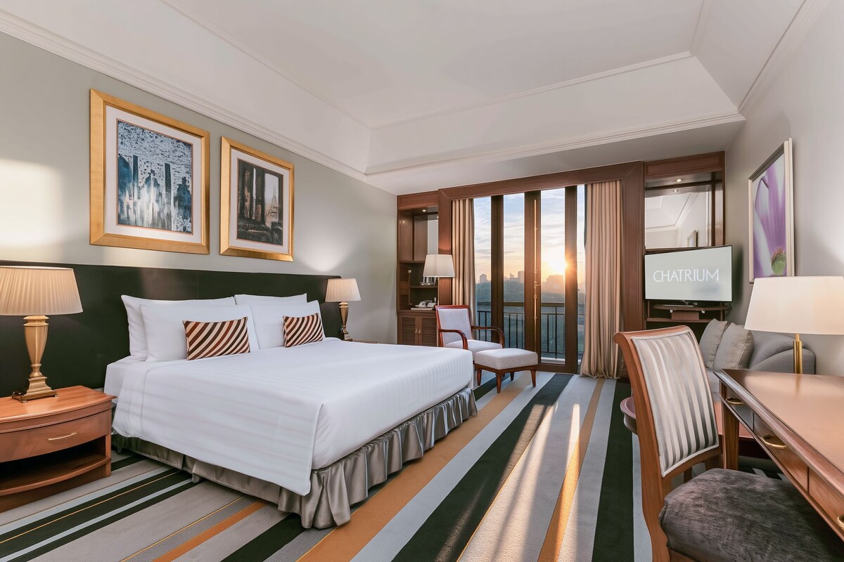 A hotel room with a large bed, two bedside tables, a TV, a seating area with a chair and a sofa, and a view of the cityscape through the window.
