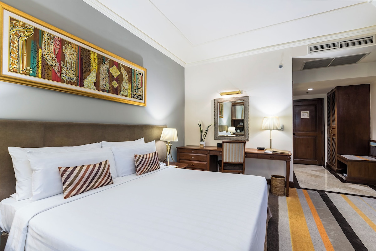 A hotel room with a large bed, a desk, a chair, a dresser, a mirror, and a decorative wall piece.