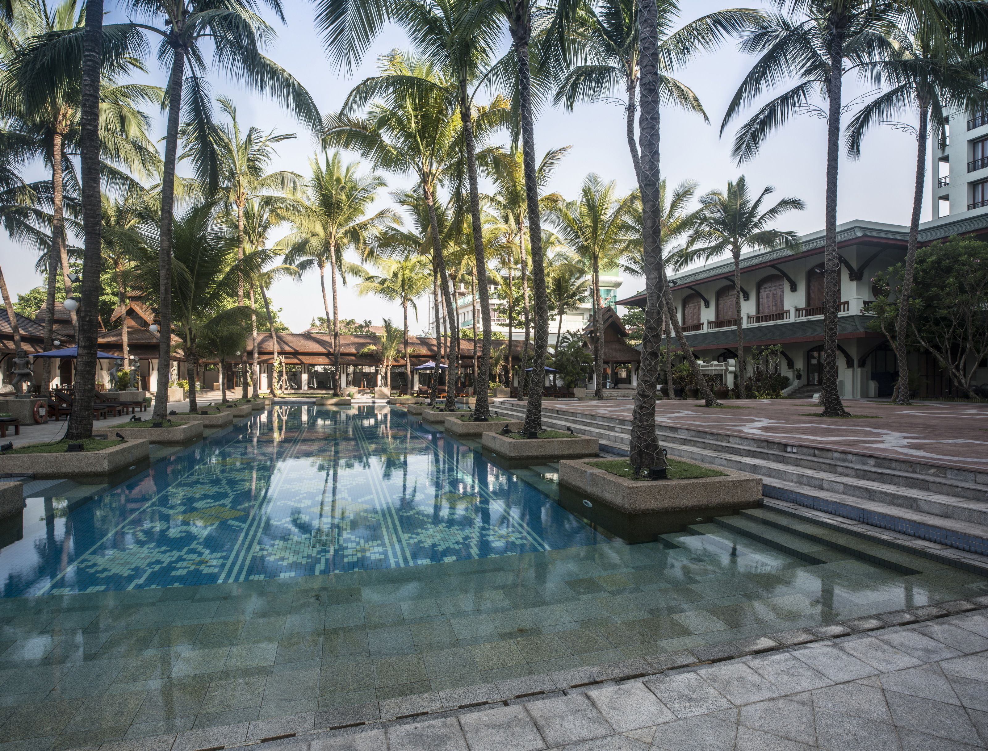 A resort pool surrounded by palm trees and buildings with a tiled walkway.