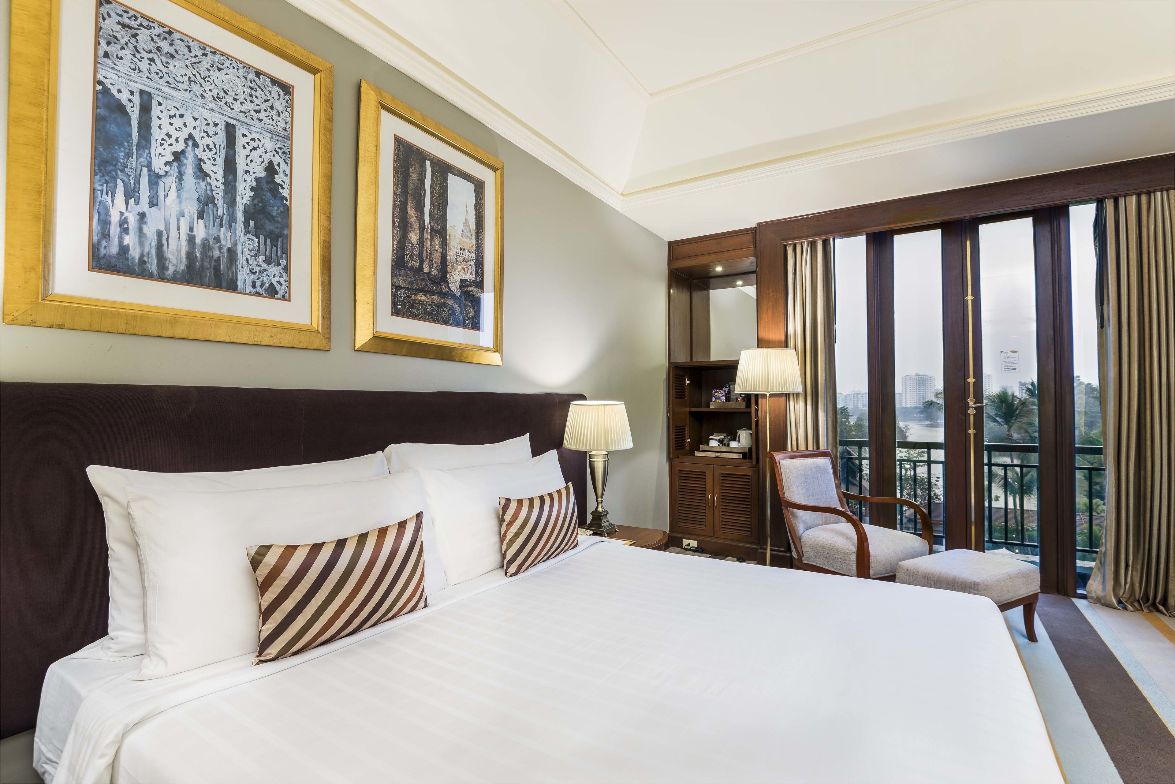 Habitación de hotel con una cama grande, dos cuadros en la pared, una mesita de noche con lámpara, una silla y vistas a la ciudad a través de amplios ventanales.