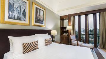 Habitación de hotel con una cama grande, dos cuadros en la pared, una mesita de noche con lámpara, una silla y vistas a la ciudad a través de amplios ventanales.