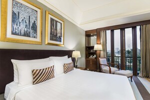 Habitación de hotel con una cama grande, dos cuadros en la pared, una mesita de noche con lámpara, una silla y vistas a la ciudad a través de amplios ventanales.