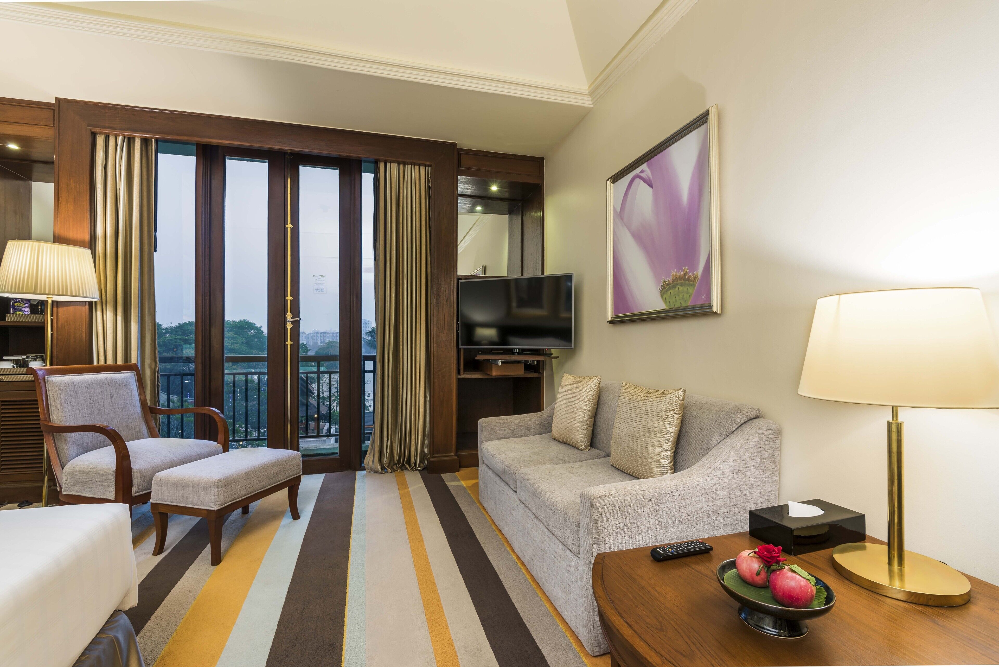 A hotel room with a sofa, armchair, TV, and a balcony with curtains.