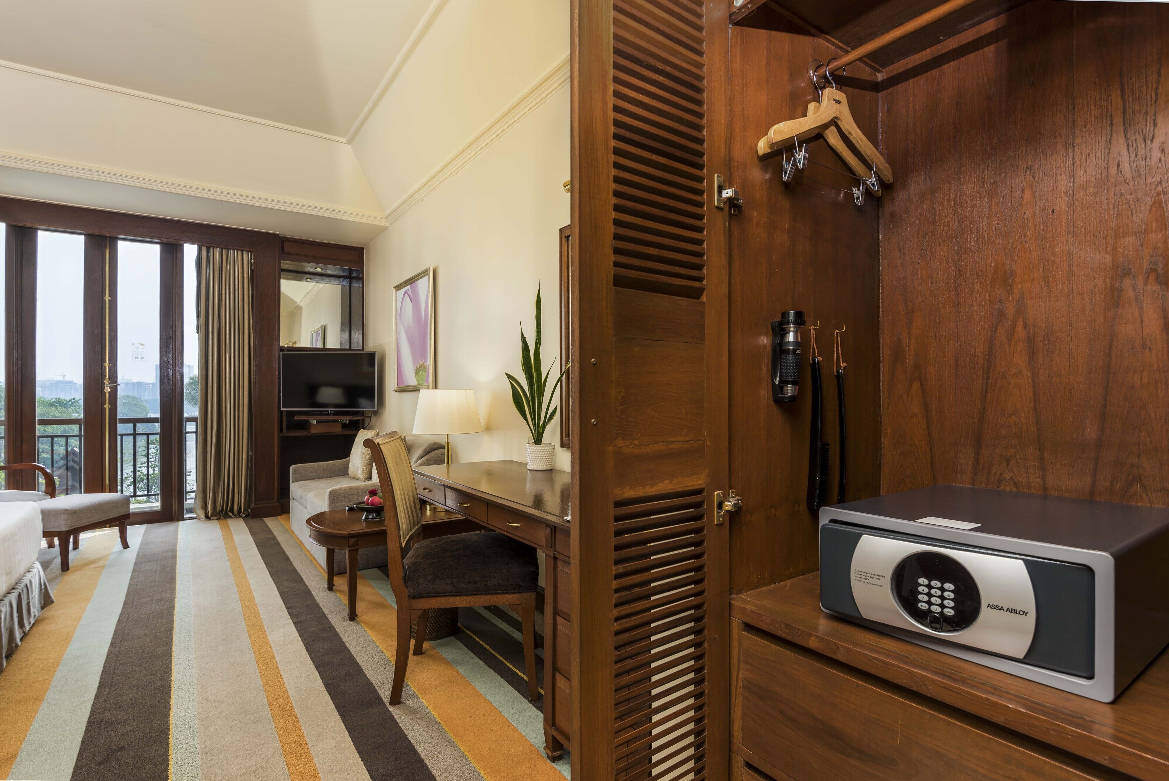 Een hotelkamer met een groot raam, een bureau, een stoel, een bank, een televisie, een lamp, een plant en een minibar.
