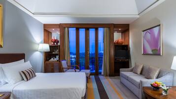 Habitación de hotel con cama, sofá, televisor, balcón con vista a la ciudad y una mesa con fruta.