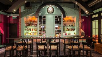 Zona de bar con un reloj, varias estanterías llenas de botellas y varios taburetes.