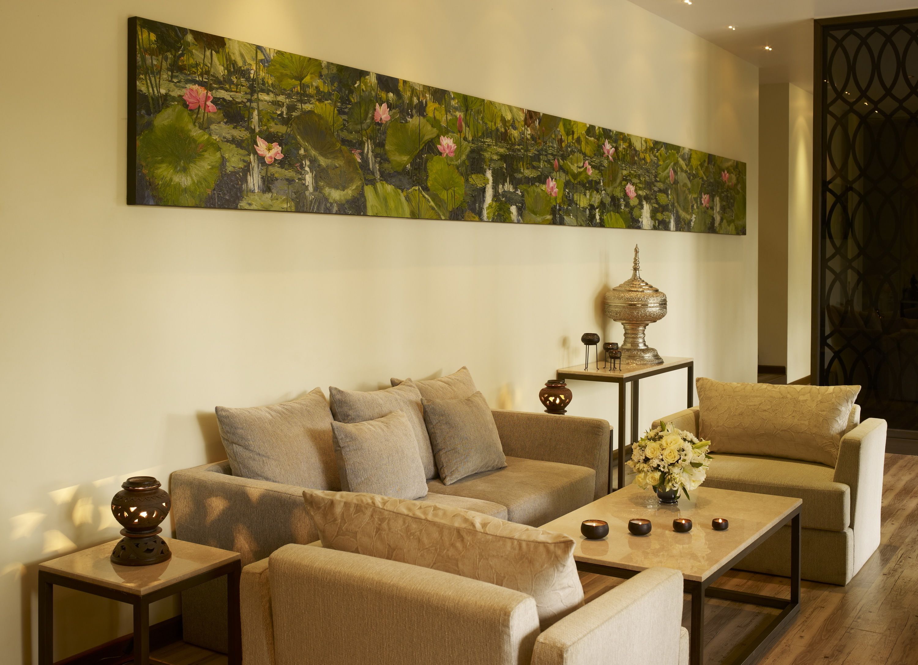 A living room with a beige sofa, a coffee table, and a large painting of a pond with lotus flowers. There is a decorative lamp and a vase with flowers on the coffee table.