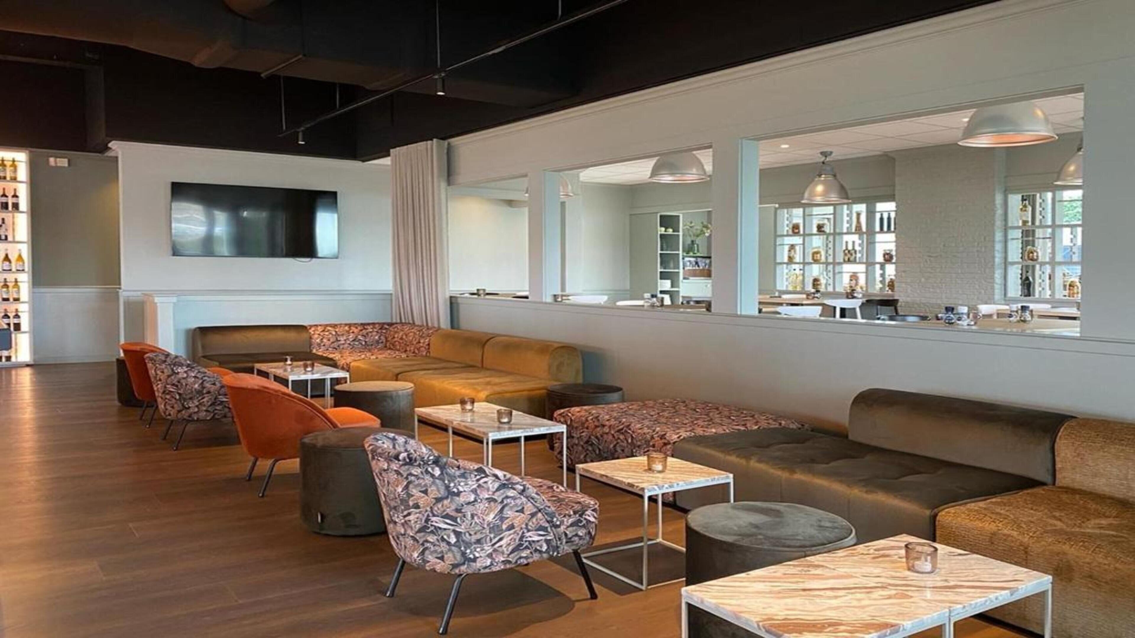 A modern lounge area with sofas, armchairs, and tables, featuring a large window with a view of a wine rack and shelves.