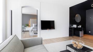 Una sala de estar moderna con un sofá gris, un televisor de pantalla plana en la pared, una pequeña mesa negra con una cesta tejida, y vista a otra habitación con un sofá blanco y una planta.