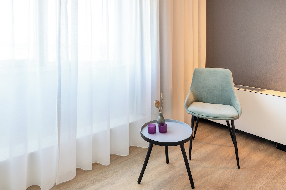 A room with a light blue armchair, a small round table with two purple candles, and a vase with a dried branch.