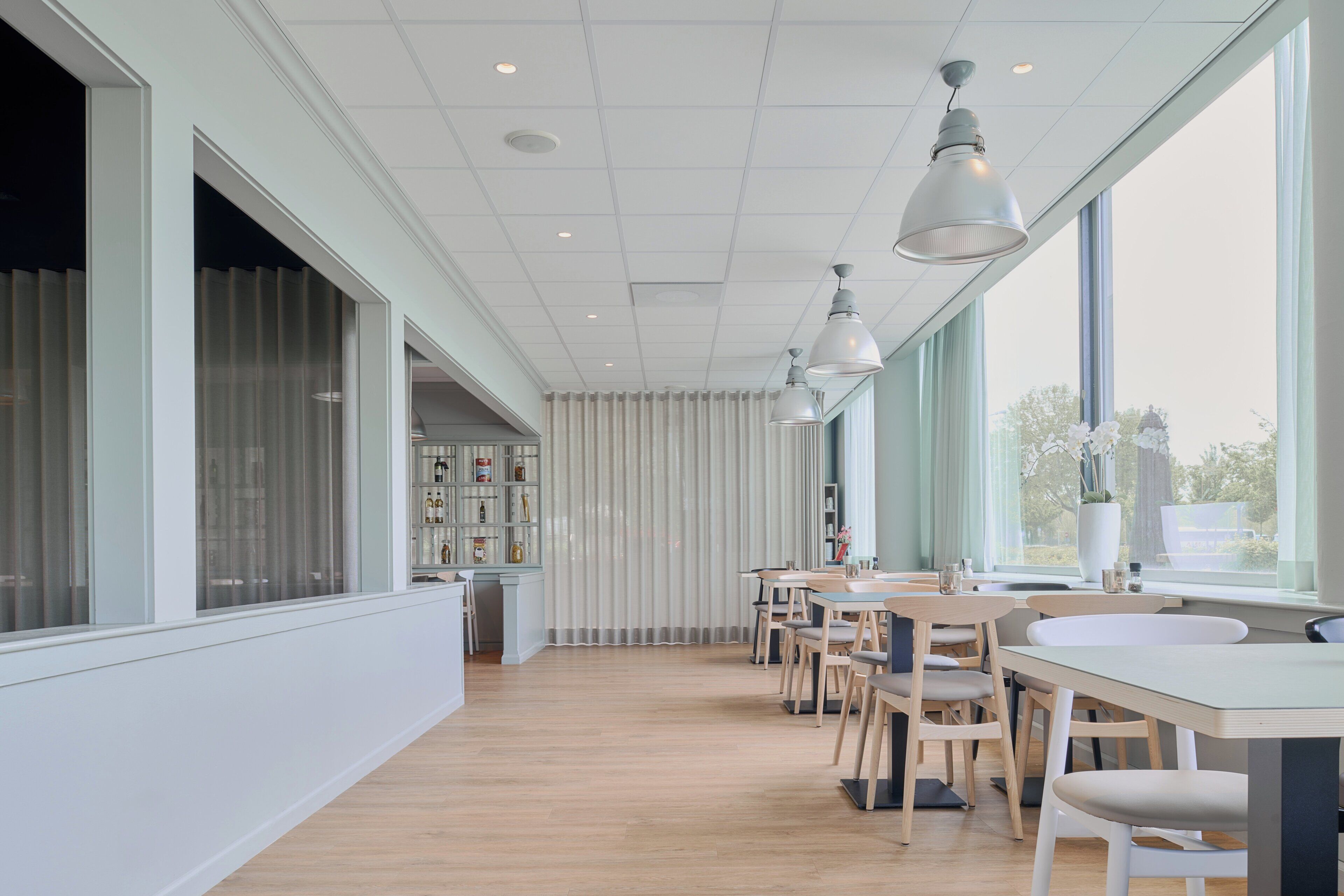 Uma sala de jantar moderna com mesas e cadeiras de madeira, janelas amplas e iluminação pendente.