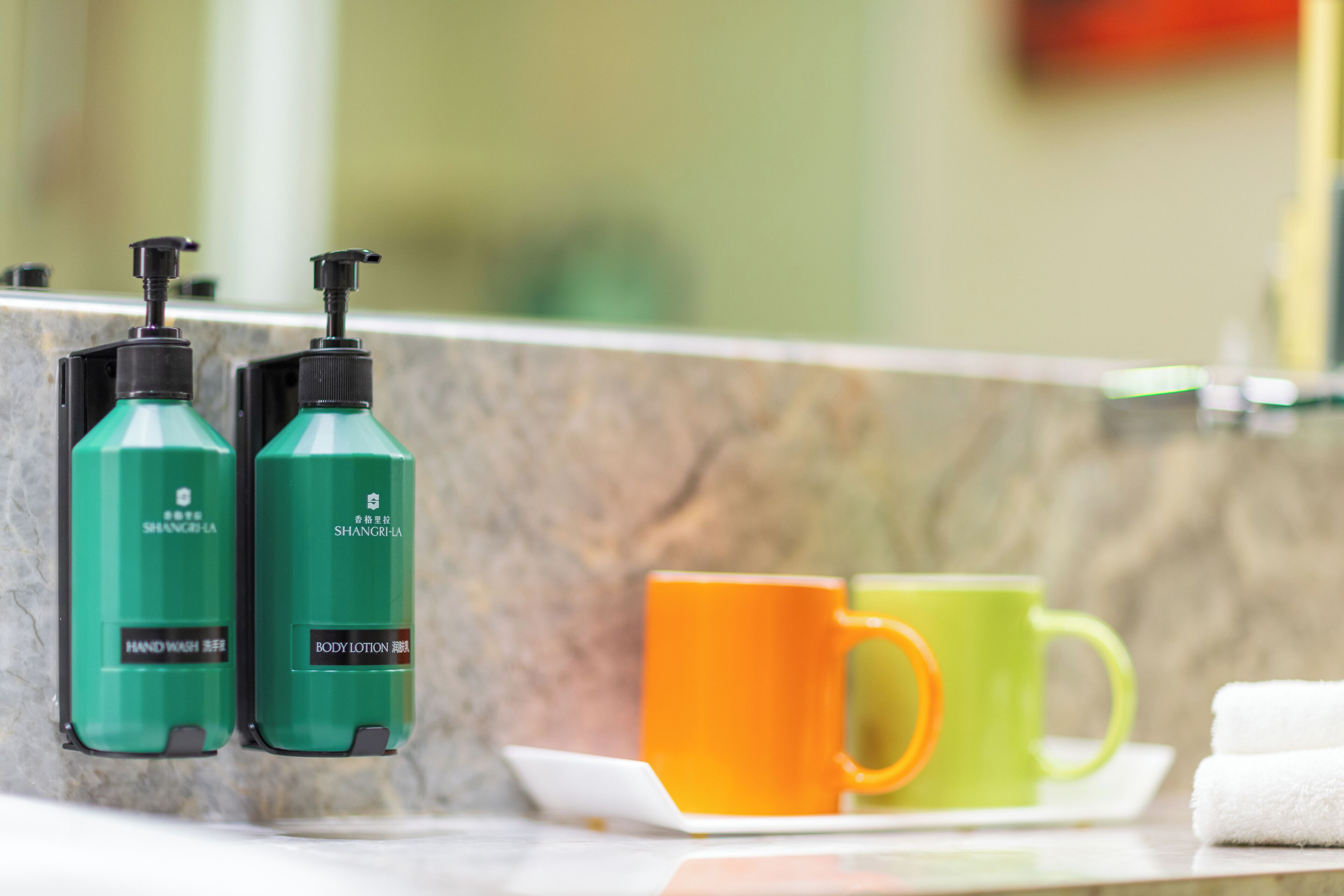 A bathroom counter with two green hand wash bottles, an orange and a green mug, and a white towel.