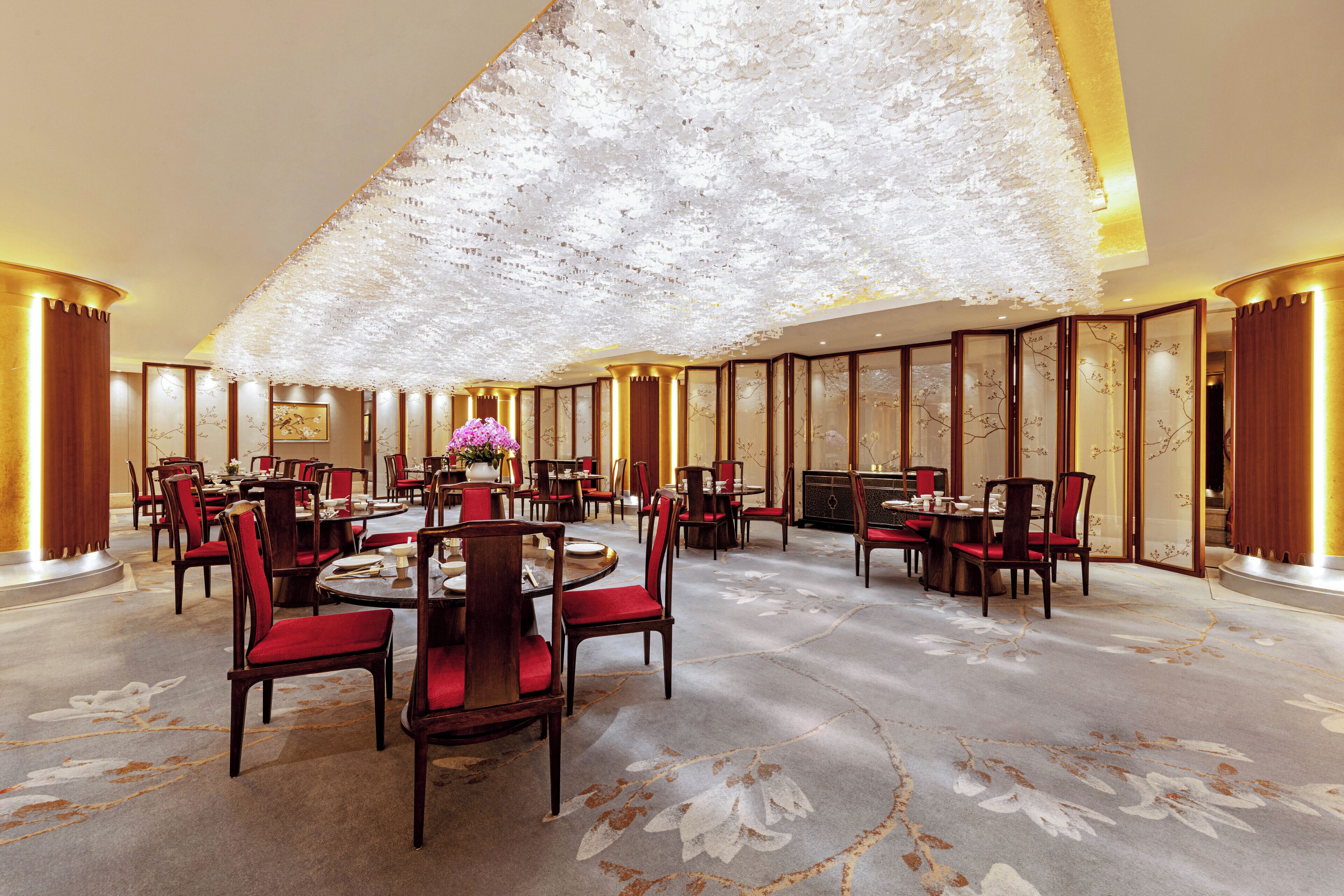 A spacious dining area with round tables and red chairs, a decorative ceiling, and traditional decor.