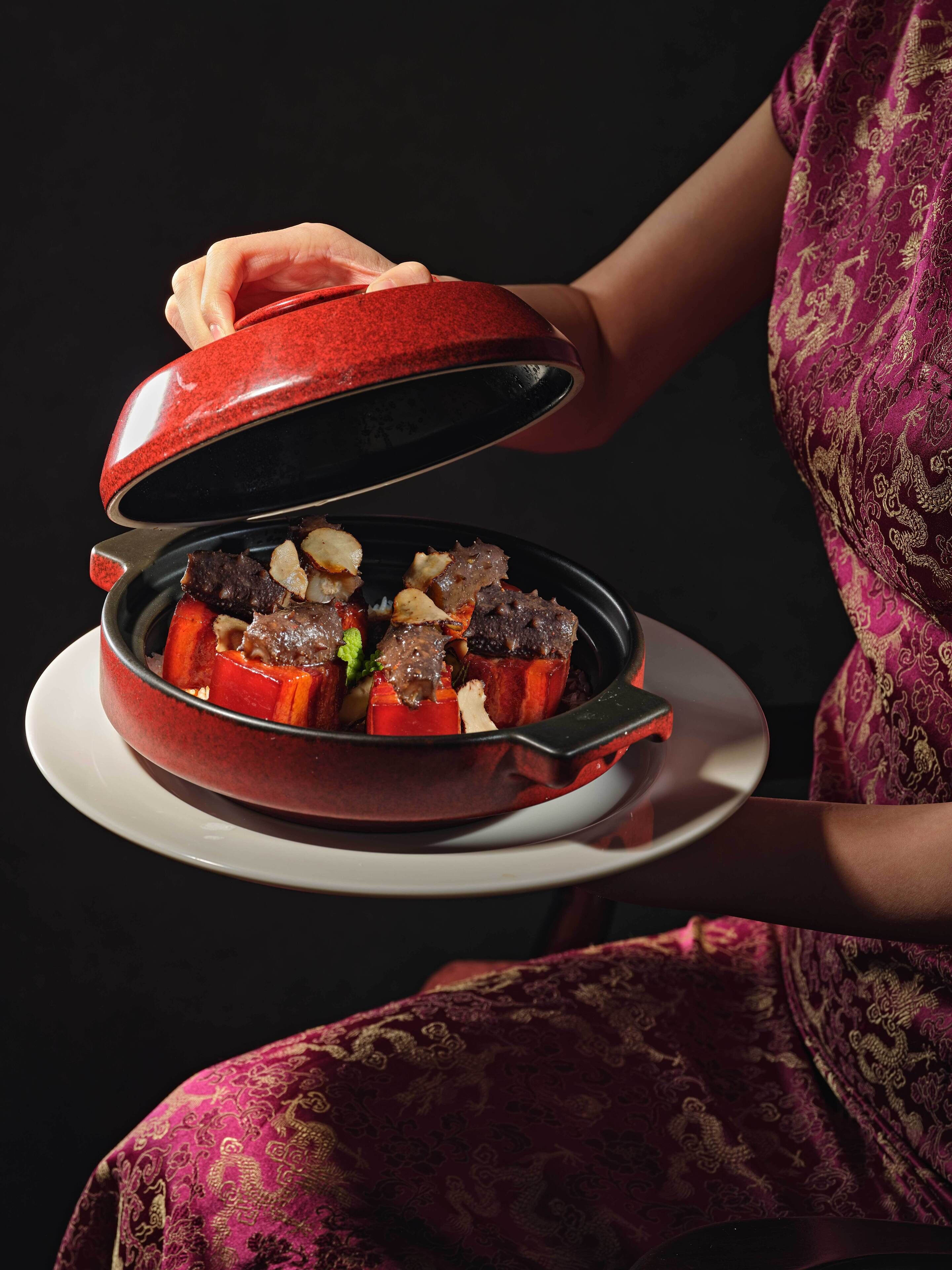 A person holding a red pot with a lid, containing a dish with vegetables and meat.