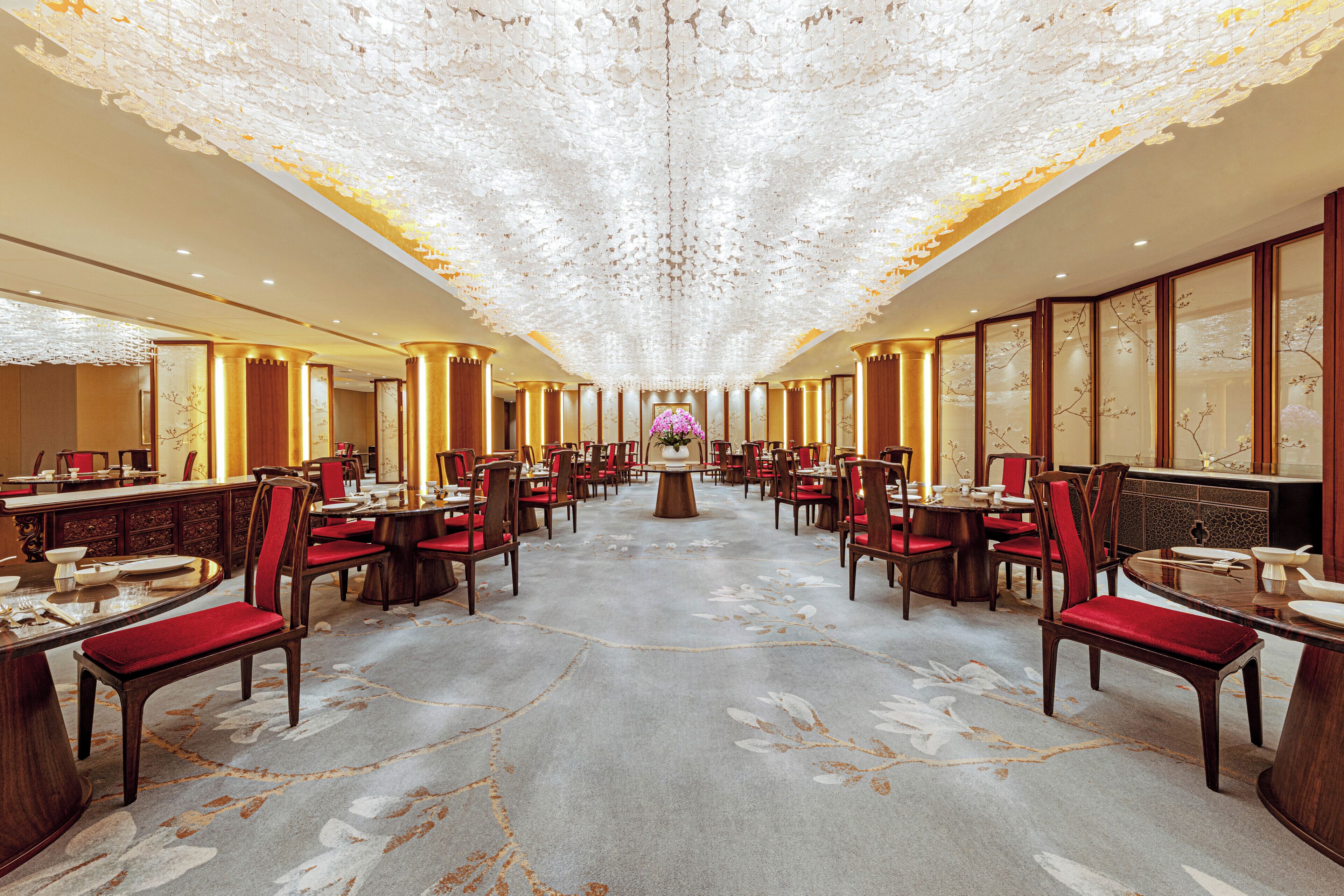 A spacious hotel lobby with a central floral arrangement, multiple dining tables, and red upholstered chairs.