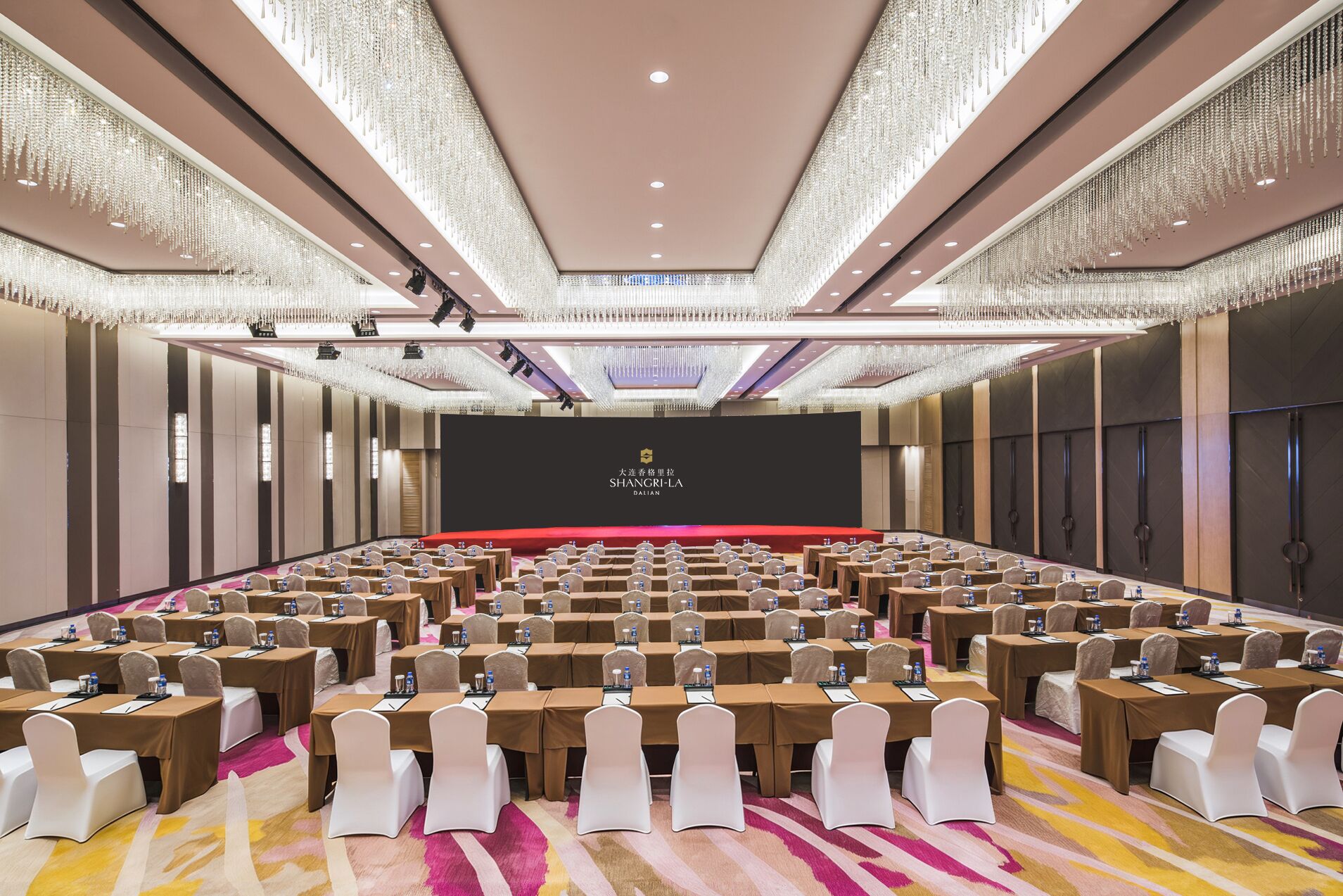 A conference room with rows of tables and chairs, a stage with a screen, and chandeliers.