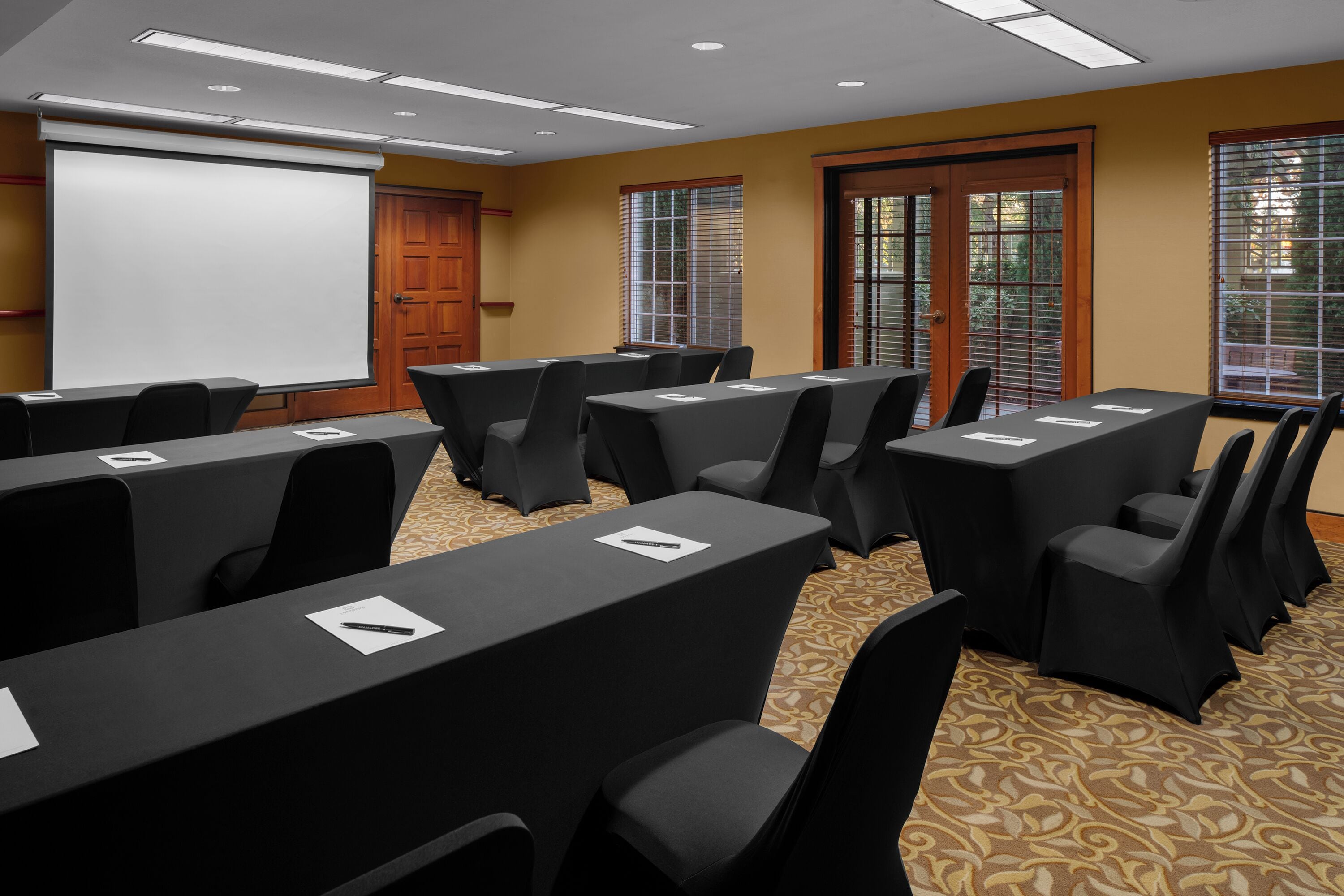 Un salón de conferencias con varias mesas y sillas, una pantalla de proyector y ventanas amplias con persianas.