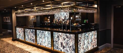 Una zona de bar con mostrador de mármol, varias botellas, vasos y una pantalla de televisión.