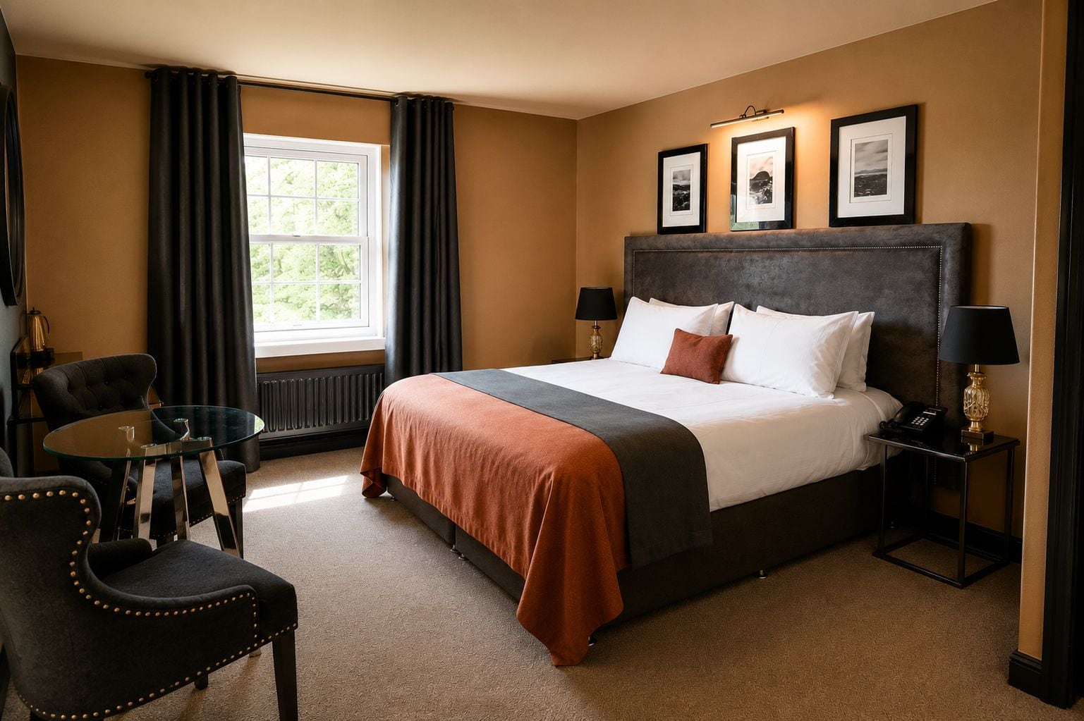 A hotel room with a large bed, a round glass table, two chairs, a radiator, and a window with curtains.