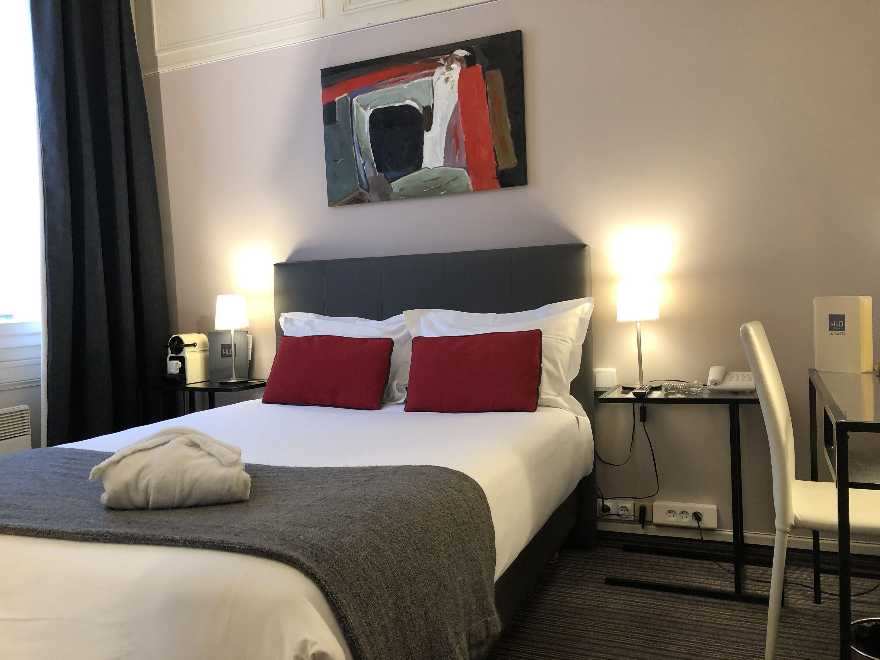 Una camera d'albergo con un letto grande, due comodini, una scrivania con una sedia, un quadro appeso al muro e una finestra con tende.
