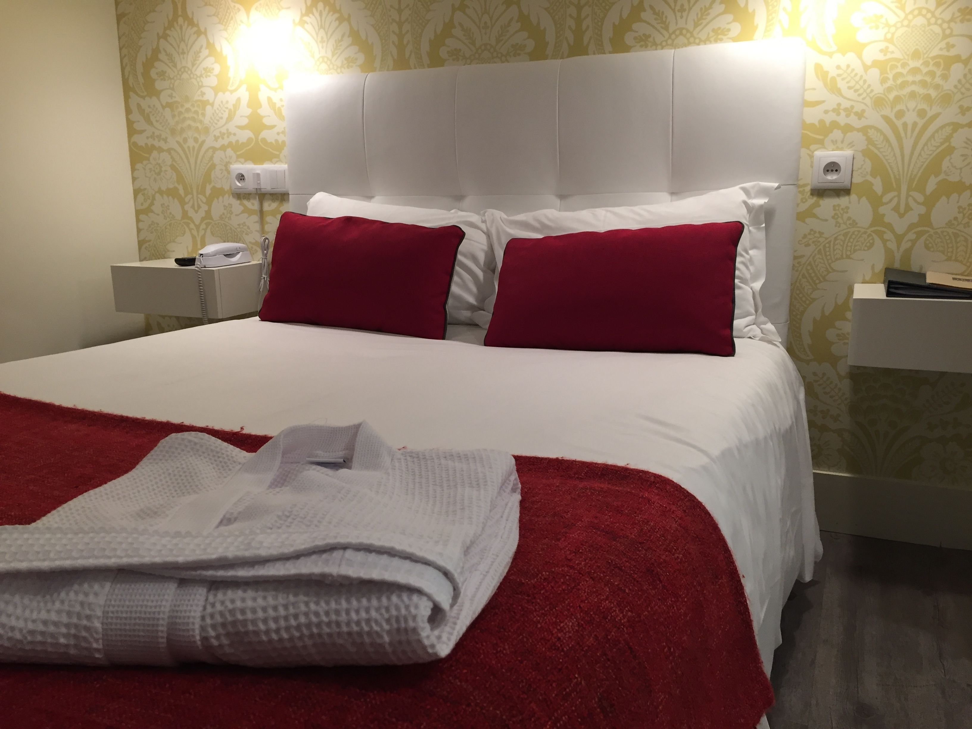 Una cama bien hecha con ropa blanca y roja, una toalla blanca doblada y una mesita de noche con un teléfono.