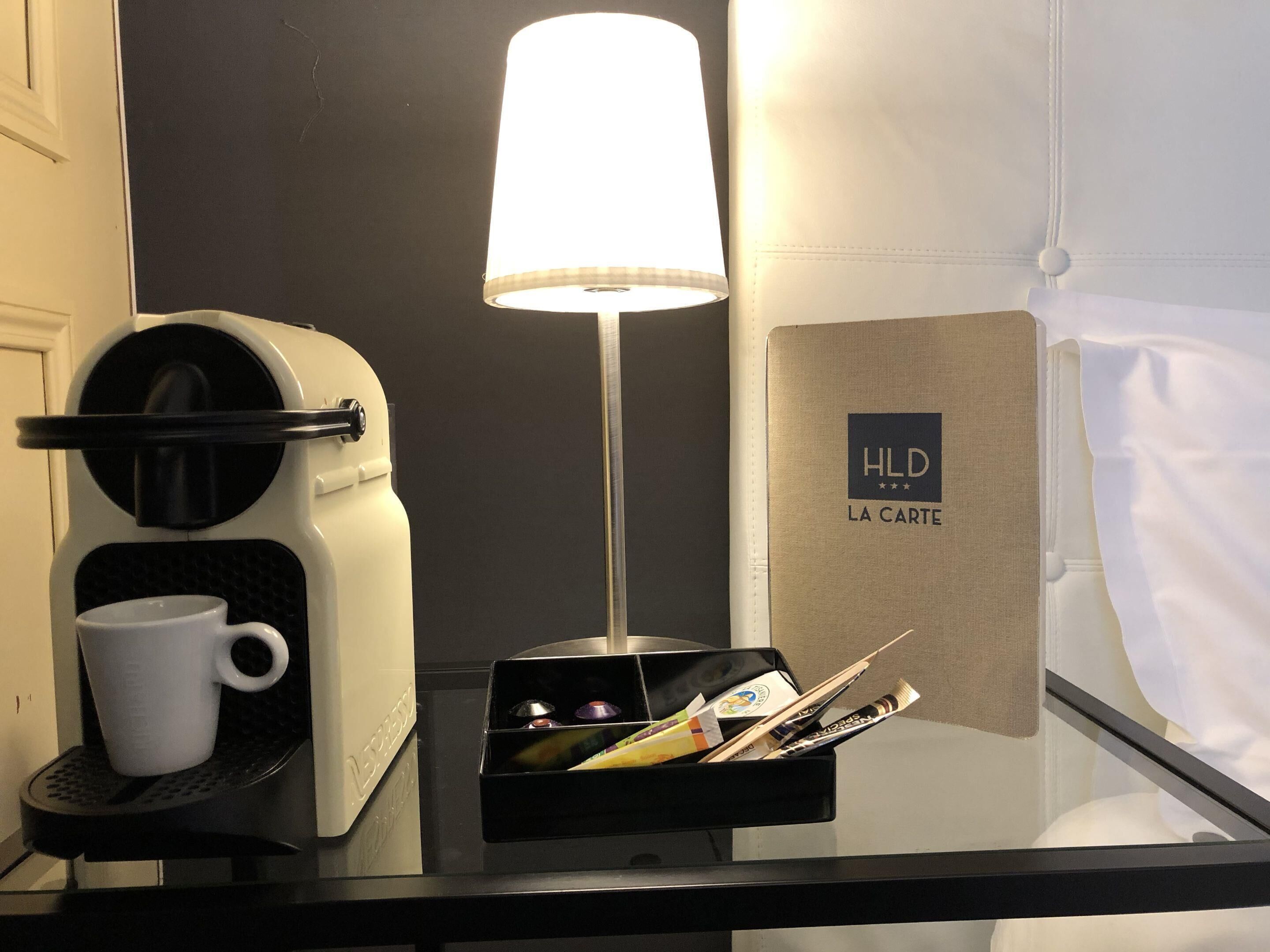 A hotel room with a coffee machine, a white mug, a tray with various items, a lamp, and a sign reading "HLD LA CARTE".