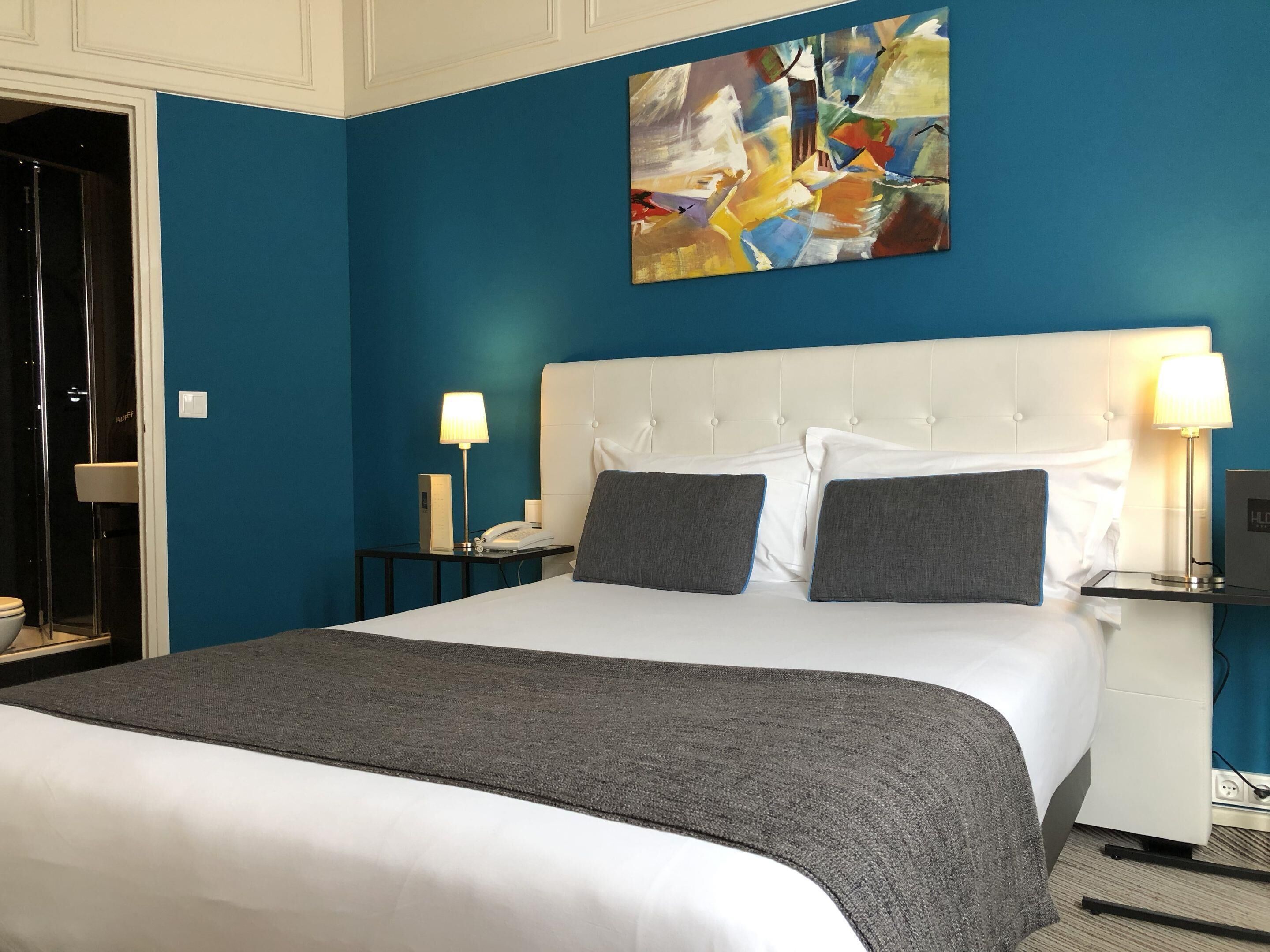 Een hotelkamer met een groot bed, een blauwe accentmuur, een modern abstract schilderij en een wit hoofdeinde.
