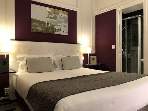 A hotel room with a large bed, two bedside tables, a painting on the wall, and a door to a bathroom. - Hôtel le Dauphin (Puteaux)