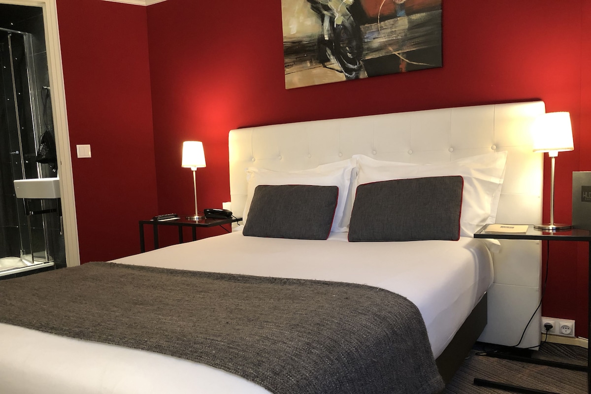 Une chambre d’hôtel avec un grand lit, deux tables de chevet avec des lampes, un mur rouge et un tableau au-dessus du lit.