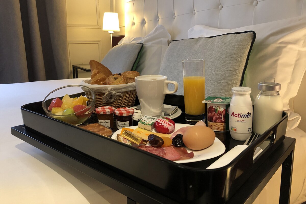 Un plateau de petit-déjeuner avec du pain, des fruits, du fromage, du jambon, des œufs, du jus et du lait, posé sur un lit dans une chambre d’hôtel.