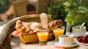 A wicker chair with a red cushion, a wicker basket with bread and a pineapple, a glass of orange juice, and a cup of coffee on a round table. - Hôtel le Dauphin (Puteaux)
