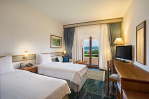 Habitación de hotel con dos camas, un escritorio, televisión y balcón con cortinas.