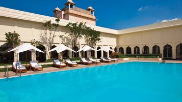 Piscina com espreguiçadeiras e guarda-sóis, um edifício com janelas em arco e uma torre.