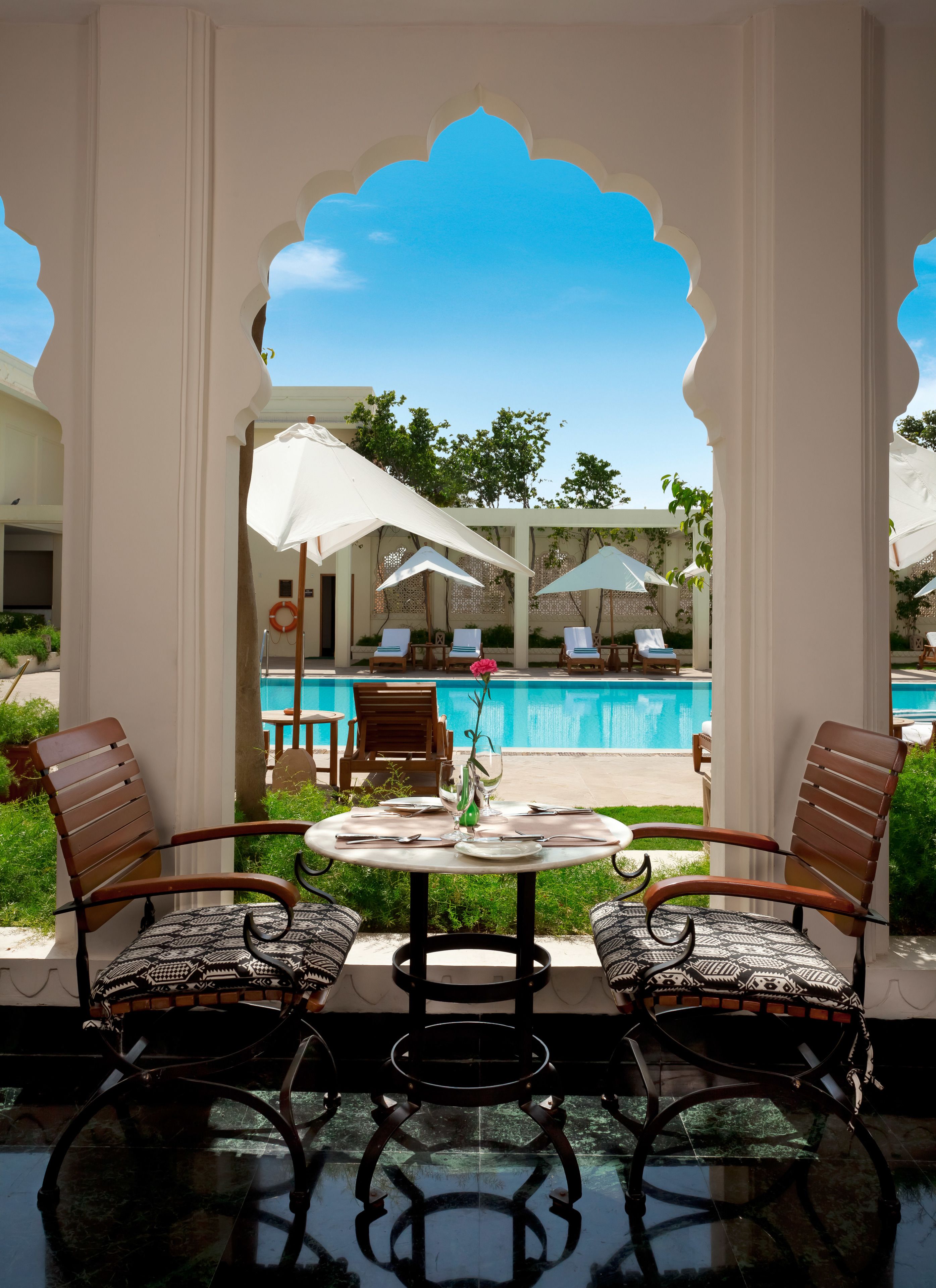 Vue depuis un patio couvert, avec deux chaises en bois et une table, donnant sur un espace piscine agrémenté de parasols et de chaises longues.