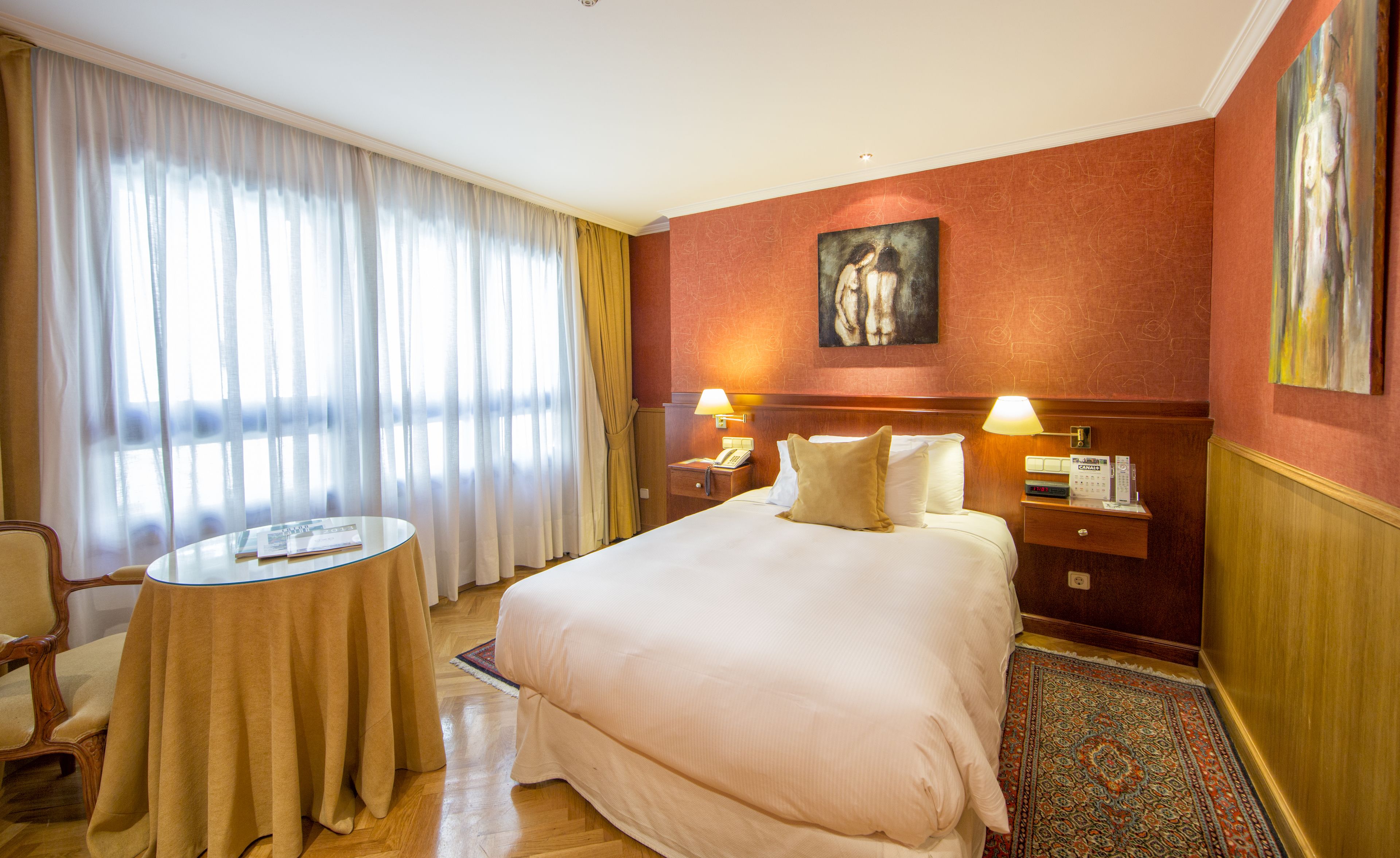 Quarto de hotel com uma cama grande, uma mesinha de cabeceira, uma cadeira, uma mesa redonda pequena e um quadro na parede.