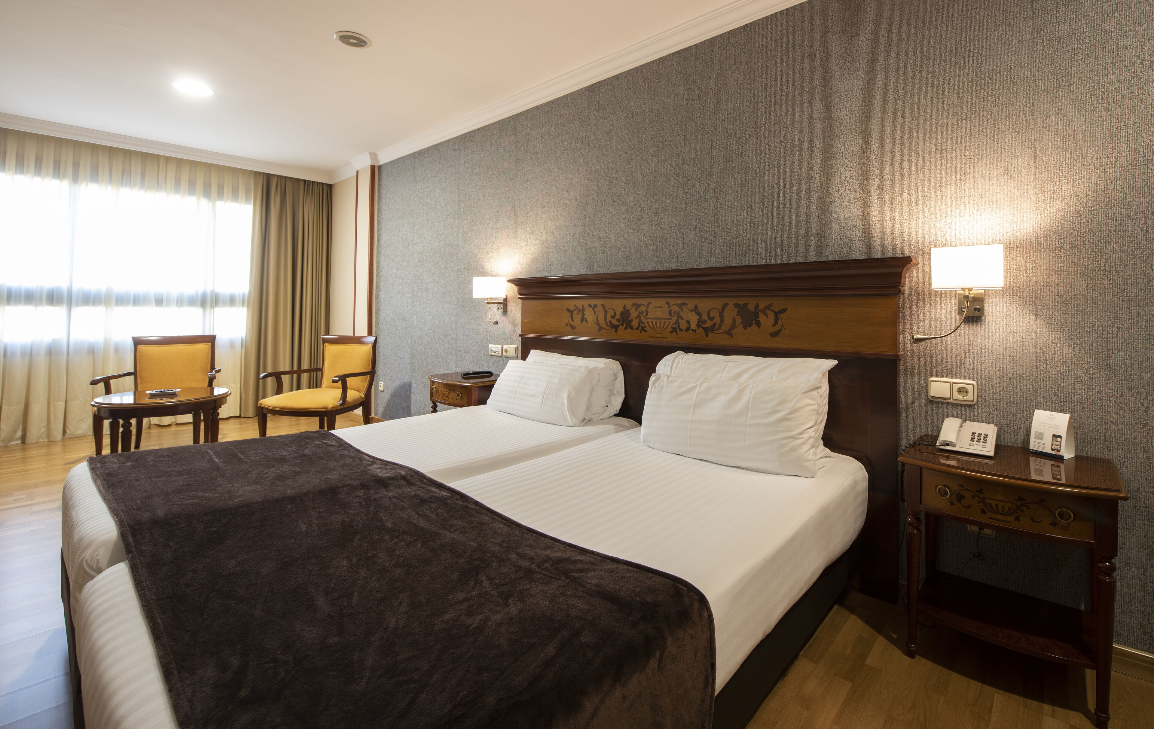 A hotel room with a large bed, two bedside tables, a nightstand, a chair, and a window with curtains.