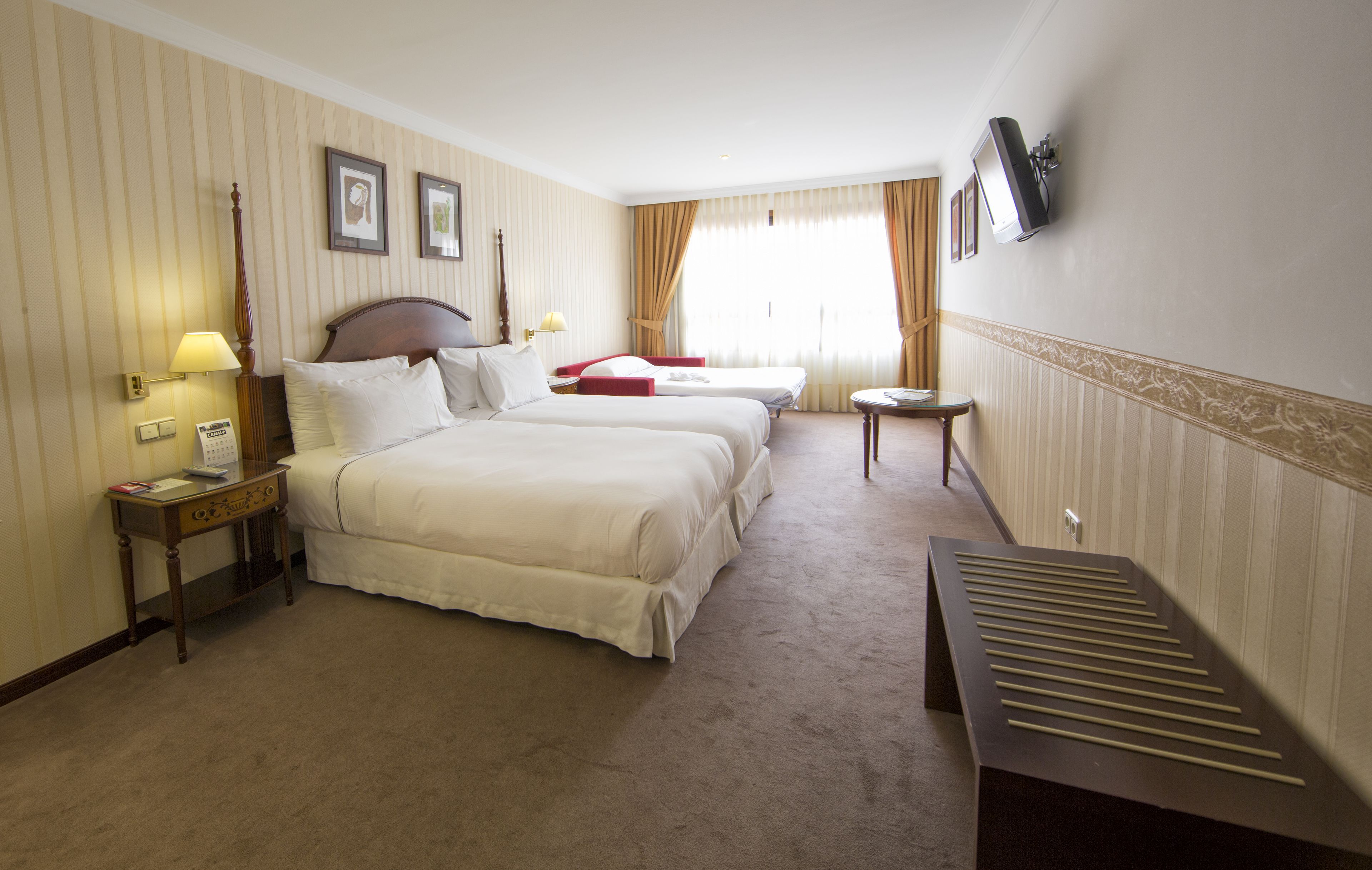 Een hotelkamer met twee bedden, een nachtkastje met een lamp, een klein rond tafeltje en een aan de muur bevestigde televisie.